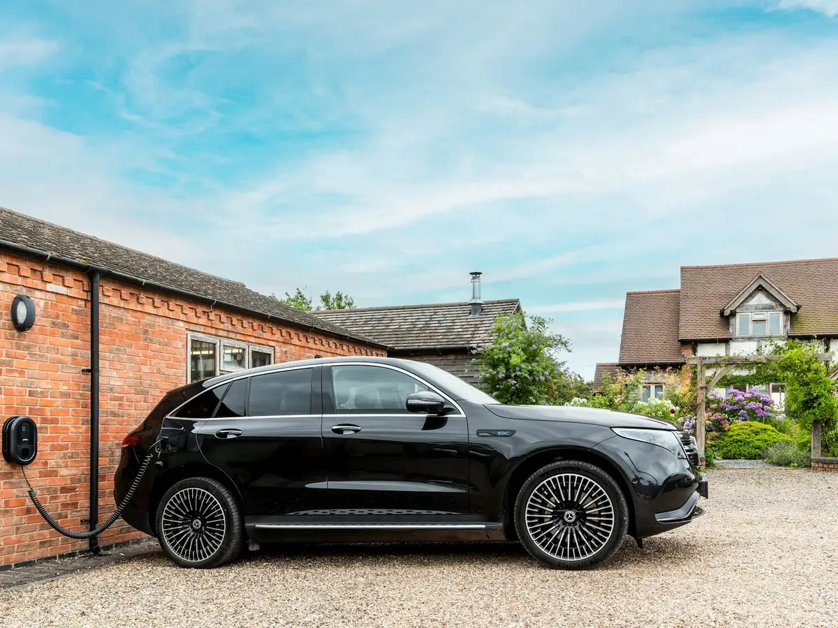 Een zwarte elektrische auto laadt aan een laadpaal op de bakstenen muur van een bijgebouw naast een huis